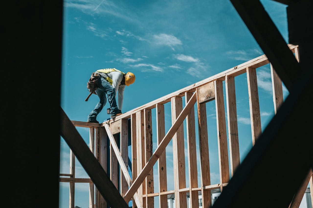 A builder in hi-vis working high on a timber house frame against a blue sky.