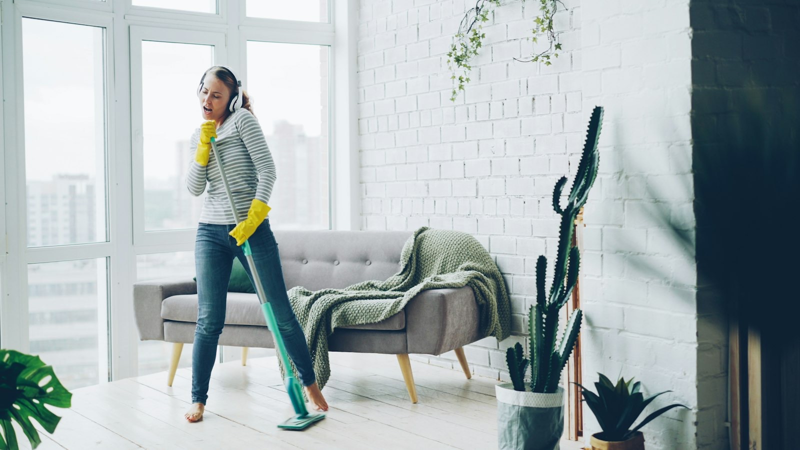 A cleaner in headphones and yellow gloves dancing with her mop in a bright living room.